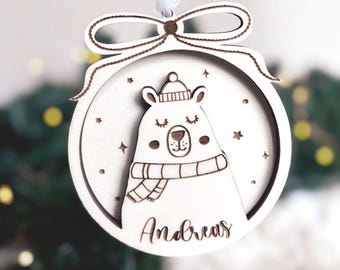 May include: White wooden Christmas ornament shaped like a circle with a polar bear wearing a scarf and hat. The ornament has the name "Andreas" written on it.