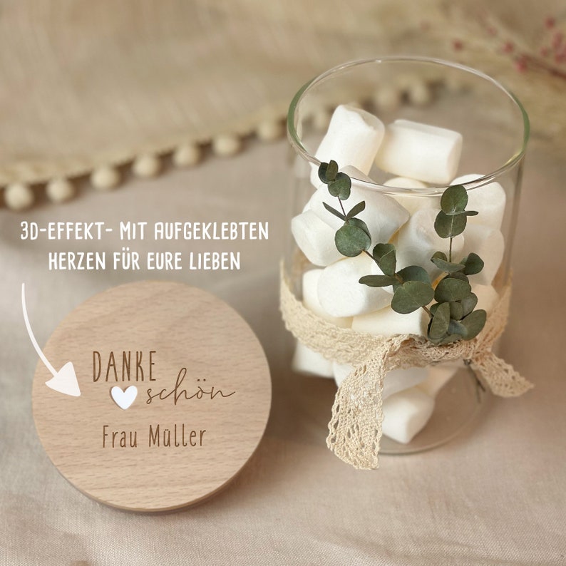 May include: A wooden round sign with the text "Danke sch&ouml;n" and a heart symbol, with the text "Frau Muller" below. The sign is next to a glass jar filled with white marshmallows and a sprig of green eucalyptus.