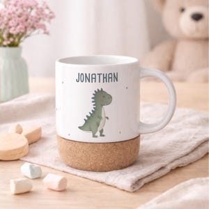 May include: White ceramic mug with a green dinosaur illustration and the name "JONATHAN" printed on it. The mug sits on a cork base. The mug has a handle and is surrounded by a soft, neutral-colored background.