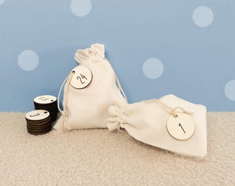 May include: Two off-white fabric drawstring pouches, each featuring a round wooden tag tied with string. One tag is marked with the number 24 and the other with the number 1. A small stack of round wooden discs with numbers is positioned to the left.