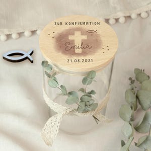 May include: A clear glass jar with a wooden lid, decorated with a cross and the name "Emilia". The lid has the text "ZUR KONFIRMATION" and the date "21.08.2021". The jar is adorned with a lace ribbon and eucalyptus.
