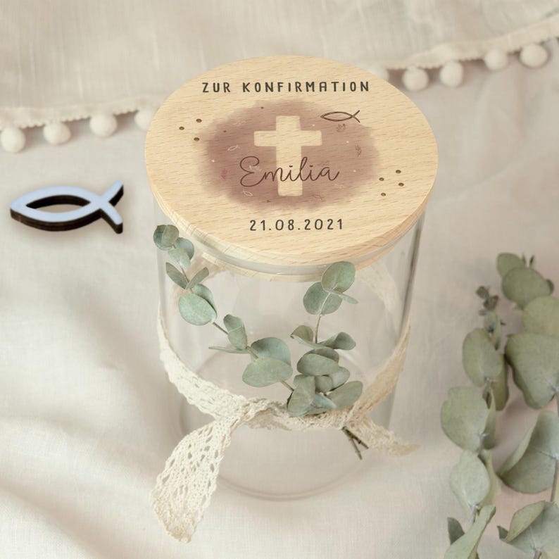 May include: A clear glass jar with a wooden lid, decorated with a cross and the name "Emilia". The lid has the text "ZUR KONFIRMATION" and the date "21.08.2021". The jar is adorned with a lace ribbon and eucalyptus.