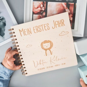 Può includere: Un album di ricordi per bambini in legno con la scritta "MEIN ERSTES JAHR" incisa sulla copertina. La copertina presenta anche un'illustrazione di leone, nuvole e il nome "Viktor Kalmár" con i dettagli della nascita: 07.10.2019, 02:33, 4050 g, 55 cm.