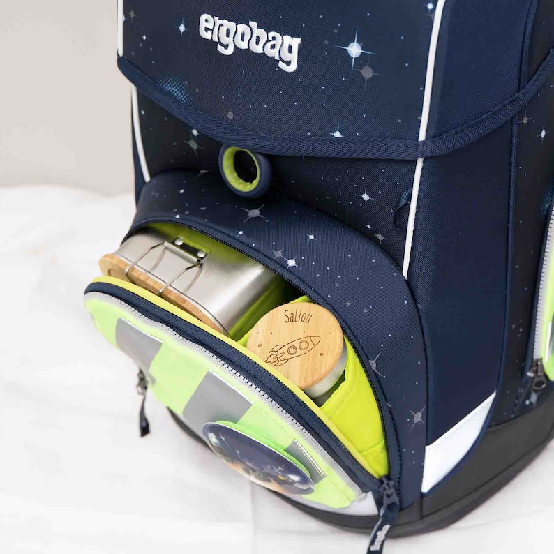 May include: A blue and green backpack with a zippered pocket. The pocket is open and contains a silver lunchbox and a wooden container with the word "Salion" written on it.