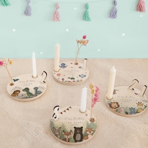 Può includere: Tre portacandele in legno con temi animali. Ogni portacandele ha uno spazio per una candela e un piccolo vaso per i fiori. I portacandele sono decorati con diversi animali, tra cui un dinosauro, un orso e un polpo. Il testo "Buon compleanno" è stampato su ogni portacandele, insieme al nome della persona che compie gli anni.