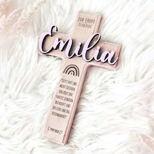 May include: Wooden cross with the name "Emilia" in pink lettering, the date "21.08.2023" and the text "Zur Taufe". The back of the cross has a rainbow design and a bible verse: "Gott hat uns nicht gegeben den Geist der Furcht, sondern der Kraft und der Liebe und der Besonnenheit" 2. Timotheus 17.