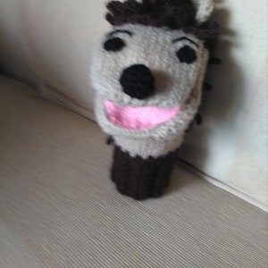 May include: A brown and grey knitted puppet with a smiling face. The puppet has a black nose and pink mouth.