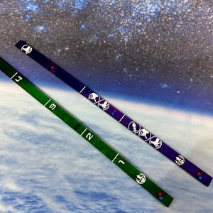 May include: Two long, thin, rectangular game pieces. One is green with white numbers and symbols, the other is purple with white symbols. Both have a series of white symbols and colored squares. The background is a space-themed image.