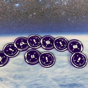 May include: A set of 11 purple game tokens with white numbers and exclamation points. The tokens are arranged in a semi-circle on a blue and white background.