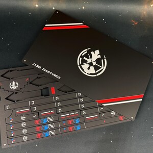 May include: A black rectangular metal sheet featuring a white circular emblem, red and white stripes, and the text LUIS MARTINEZ. The sheet contains various laser-cut components, including numbered strips, small geometric shapes, and blue and red icons.