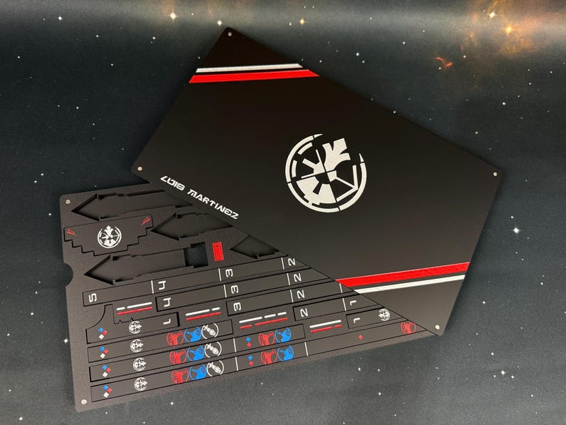May include: A black rectangular metal sheet featuring a white circular emblem, red and white stripes, and the text LUIS MARTINEZ. The sheet contains various laser-cut components, including numbered strips, small geometric shapes, and blue and red icons.