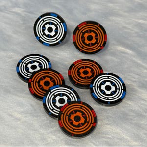 Puede incluir: Una colección de fichas de juego circulares negras con diseños de círculos concéntricos. Algunas fichas tienen detalles blancos y azules, mientras que otras presentan detalles naranjas y rojos. Las fichas están dispuestas sobre una superficie gris claro.