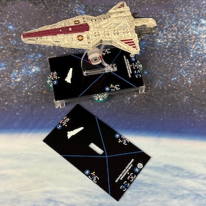 Puede incluir: Modelo de una nave espacial Star Destroyer, de color crema y burdeos, expuesto sobre dos cartas de juego negras. Las cartas tienen siluetas de barcos blancos y símbolos de juego. El fondo es una escena espacial.
