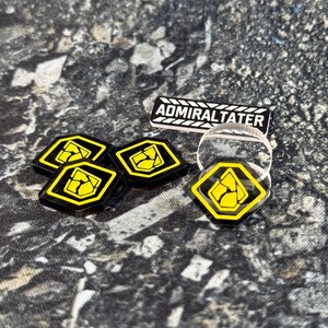 May include: A set of four black and yellow hexagonal tokens featuring a stylised cracked stone icon, accompanied by a rectangular black and white nameplate with the text ADMIRALTATER in a bold, sans-serif font, and a clear circular acrylic ring.
