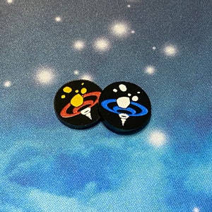 May include: Two black circular game pieces with white, blue, and red painted designs. The designs are stylized depictions of a planet with rings and a star.