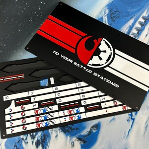 May include: A rectangular black metal game board with a lid featuring a red and white graphic design, the text "TO YOUR BATTLE STATIONS!", and symbols of two opposing factions. The base contains recessed slots and white printed grids with icons.