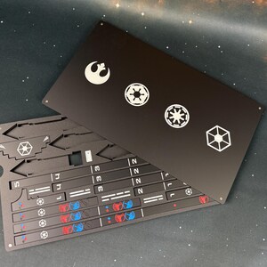 May include: A black rectangular metal sheet featuring white laser-etched Star Wars symbols including the Rebel Alliance logo and Imperial emblems. Beneath it lies a matching black metal component sheet with pre-cut shapes, numbered sections, and small red and blue circular icons.
