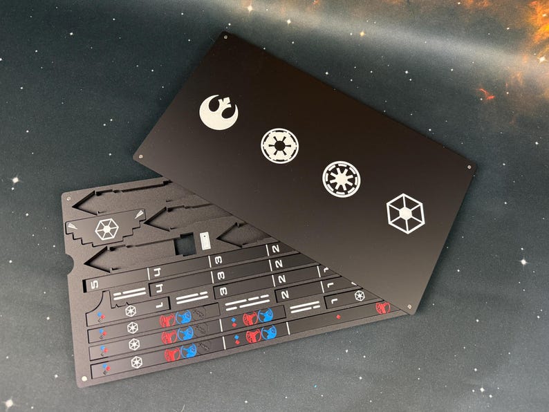 May include: A black rectangular metal sheet featuring white laser-etched Star Wars symbols including the Rebel Alliance logo and Imperial emblems. Beneath it lies a matching black metal component sheet with pre-cut shapes, numbered sections, and small red and blue circular icons.