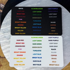 May include: Two rectangular color sample cards, one black and one white, each featuring three columns of printed text labels. The text lists various color names in their corresponding hues, including white, red, orange, yellow, green, blue, purple, gray, and metallic tones like silver, copper, brass, and gold.