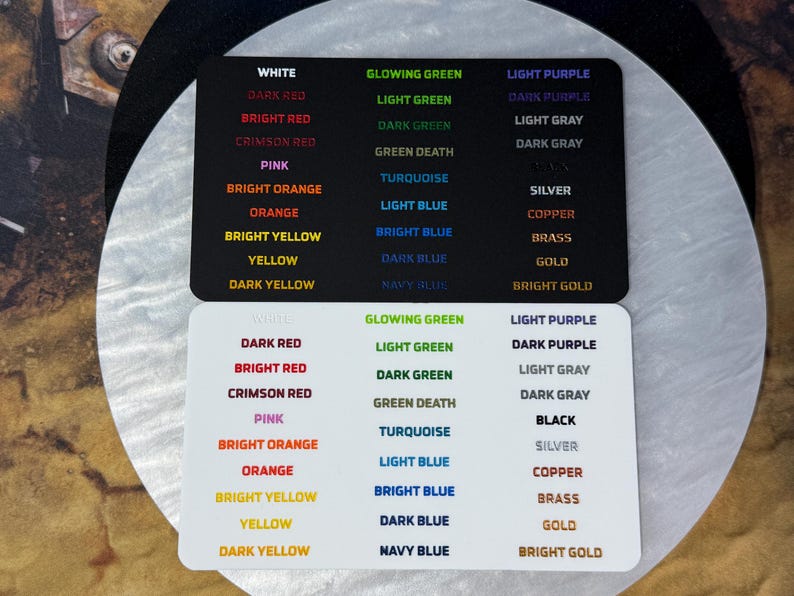 May include: Two rectangular color sample cards, one black and one white, each featuring three columns of printed text labels. The text lists various color names in their corresponding hues, including white, red, orange, yellow, green, blue, purple, gray, and metallic tones like silver, copper, brass, and gold.