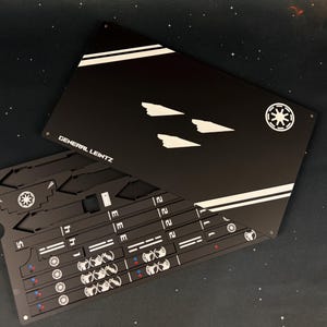 May include: A black rectangular metal sheet featuring white graphic designs, including three ship silhouettes, two diagonal stripes, a circular emblem, and the text GENERAL LEINTZ. Beneath it, a second black sheet displays various cut-out shapes.