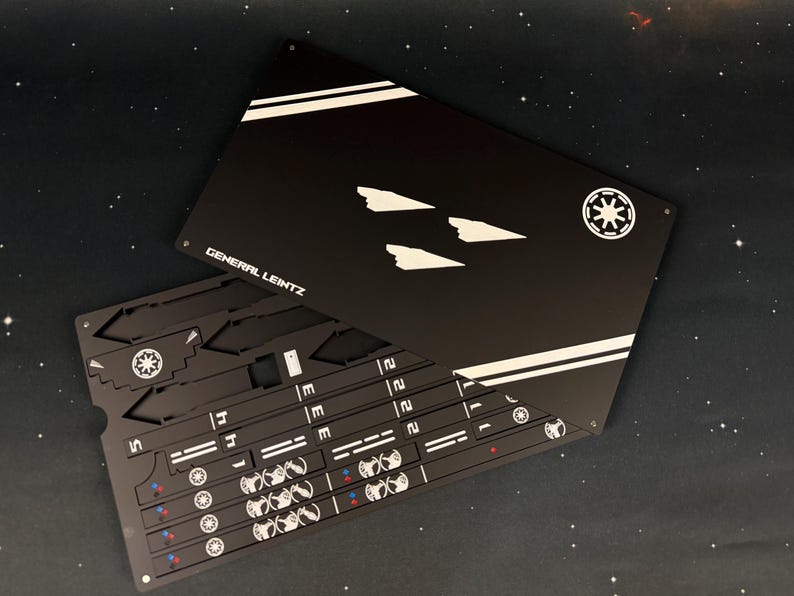 May include: A black rectangular metal sheet featuring white graphic designs, including three ship silhouettes, two diagonal stripes, a circular emblem, and the text GENERAL LEINTZ. Beneath it, a second black sheet displays various cut-out shapes.