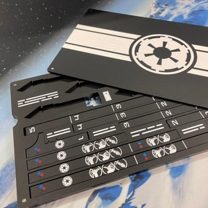 May include: A black rectangular game dashboard with a removable lid featuring a white Galactic Empire symbol. The base contains recessed slots, numbered sections, and white printed icons including small red and blue diamond shapes and circular emblems.