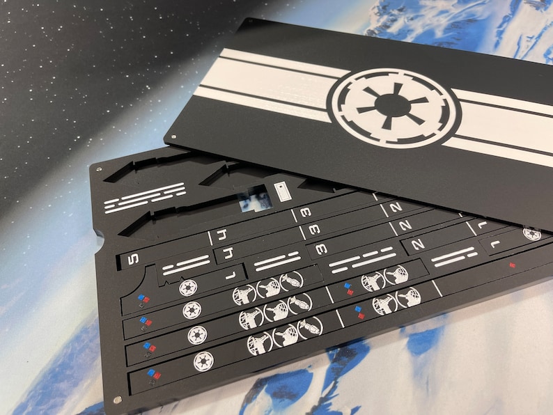May include: A black rectangular game dashboard with a removable lid featuring a white Galactic Empire symbol. The base contains recessed slots, numbered sections, and white printed icons including small red and blue diamond shapes and circular emblems.