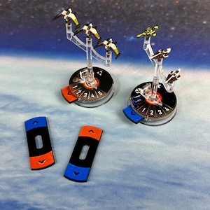 Könnte beinhalten: Drei Miniatur-Raumschiffe auf durchsichtigen Plastikständern mit schwarz-weißen nummerierten Basen. Zwei orangefarbene und blaue Manövrier-Zifferblätter auf blau-weißem Hintergrund.