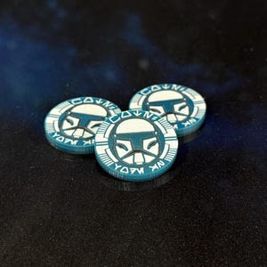 May include: Three teal and white circular tokens with a helmet design. The tokens have a white border with text and a teal centre featuring a stylized helmet. The tokens are arranged on a dark, reflective surface.