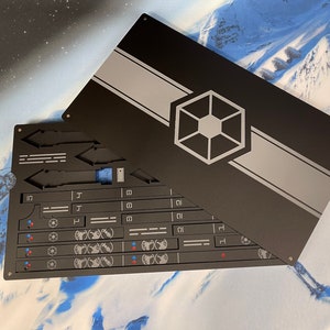 May include: A rectangular black metal case with a sliding lid featuring a grey horizontal stripe and a central hexagonal emblem. The interior contains organized slots for various flat components, including small rectangular pieces with white symbols.