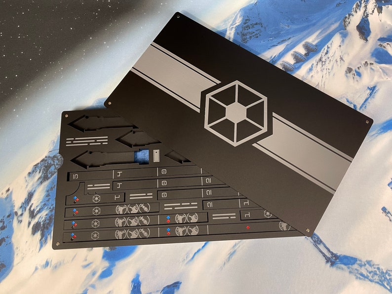 May include: A rectangular black metal case with a sliding lid featuring a grey horizontal stripe and a central hexagonal emblem. The interior contains organized slots for various flat components, including small rectangular pieces with white symbols.