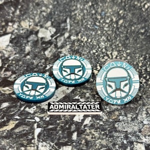 May include: Two circular teal and white tokens featuring a stylized clone trooper helmet graphic and Aurebesh text, alongside a larger circular token with the same design and a rectangular black and white plaque with the text ADMIRALTATER.