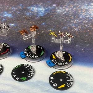 Puede incluir: Miniaturas de cazas estelares de varios colores, montadas sobre soportes transparentes, con bases negras que muestran nombres como "VADER" y "ANAKIN". Las bases tienen indicadores direccionales y marcas numéricas. El fondo es un diseño temático espacial.