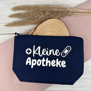 Könnte beinhalten: Eine marineblaue Reißverschlusstasche mit den Worten "Kleine Apotheke" und einer Bandagen-Grafik in Weiß. Die Tasche ist rechteckig und liegt auf einem rosa Waffelstoff. Getrocknete Pflanzen und eine Holzscheibe befinden sich im Hintergrund.