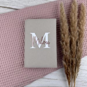 Könnte beinhalten: Ein beigefarbener Reisepasshalter mit einem großen weißen Großbuchstaben M, über den der Name Milou in einer roségoldenen Schreibschrift gelegt ist. Der Einband hat eine strukturierte Oberfläche und eine rechteckige Form.