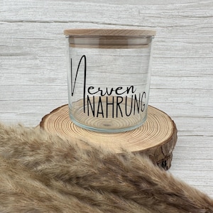 Könnte beinhalten: Ein klares Glas mit einem Holzdeckel, auf dem in schwarzer Schrift die Worte "Nerven Nahrung" stehen. Das Glas steht auf einer Holzscheibe, im Vordergrund getrocknetes Ziergras. Das Glas ist leer.