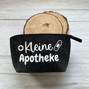 Könnte beinhalten: Eine kleine, dunkelgraue Reißverschlusstasche mit dem weißen Schriftzug "Kleine Apotheke", einem Kreuz und einer Bandagen-Grafik. Die Tasche befindet sich vor einem Holzuntergrund.