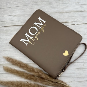 Könnte beinhalten: Eine braune Mom Organizer Clutch mit goldenem Reißverschluss und Handschlaufe. Die Worte "MOM Organizer" sind in Weiß und Gold gedruckt. Auf der Vorderseite der Clutch befindet sich ein goldenes Herz.