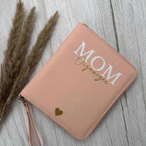 Könnte beinhalten: Ein rosafarbener Organizer mit dem Wort "MOM" in weißen Blockbuchstaben und "Organizer" in goldener Schrift. Ein goldenes Herz befindet sich in der unteren linken Ecke. Der Organizer hat einen Reißverschluss und eine Handschlaufe.