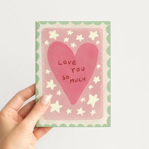 May include: A rectangular greeting card featuring a large pink heart in the centre with the text "LOVE YOU SO MUCH" written in dark red. The background is light pink with scattered cream-coloured stars and a scalloped light green border.