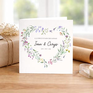 May include: A square white wedding card featuring a heart-shaped floral wreath. The wreath is made of green foliage and purple, pink, and yellow flowers. The card displays the names "Ioan & Cerys" and the date "20-03-26".