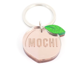 May include: A rose gold peach-shaped keychain with the word "MOCHI" engraved on it. The keychain has a green glitter leaf charm attached to it.
