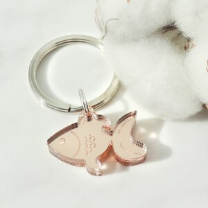 Puede incluir: Un llavero de pez de color oro rosa con un anillo de llave plateado. El pez tiene un dise&ntilde;o detallado con escamas y una aleta.