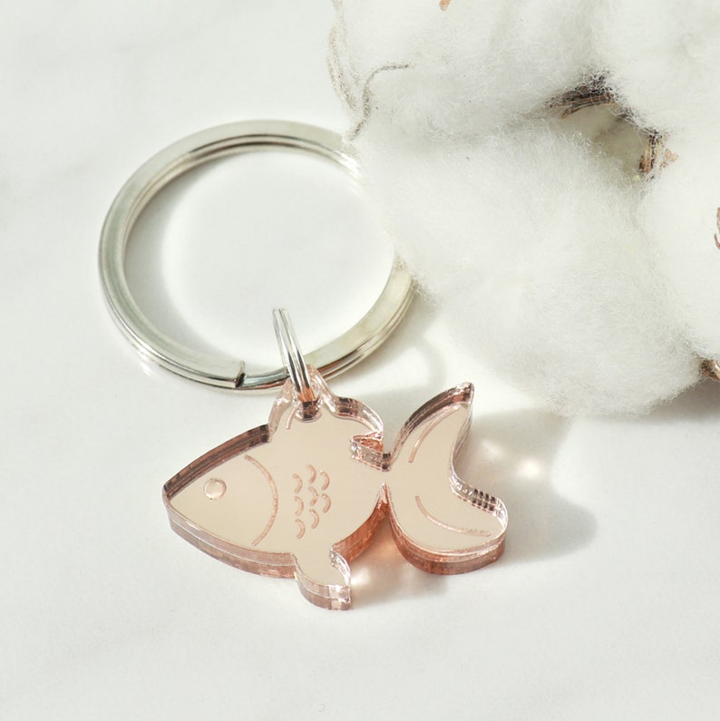 Puede incluir: Un llavero de pez de color oro rosa con un anillo de llave plateado. El pez tiene un dise&ntilde;o detallado con escamas y una aleta.