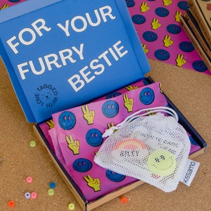 Puede incluir: Una caja azul con el texto "FOR YOUR FURRY BESTIE" y una peque&ntilde;a bolsa blanca con una carita sonriente amarilla en su interior. La caja est&aacute; abierta y el interior est&aacute; forrado con papel rosa con caritas sonrientes azules y signos de paz amarillos.