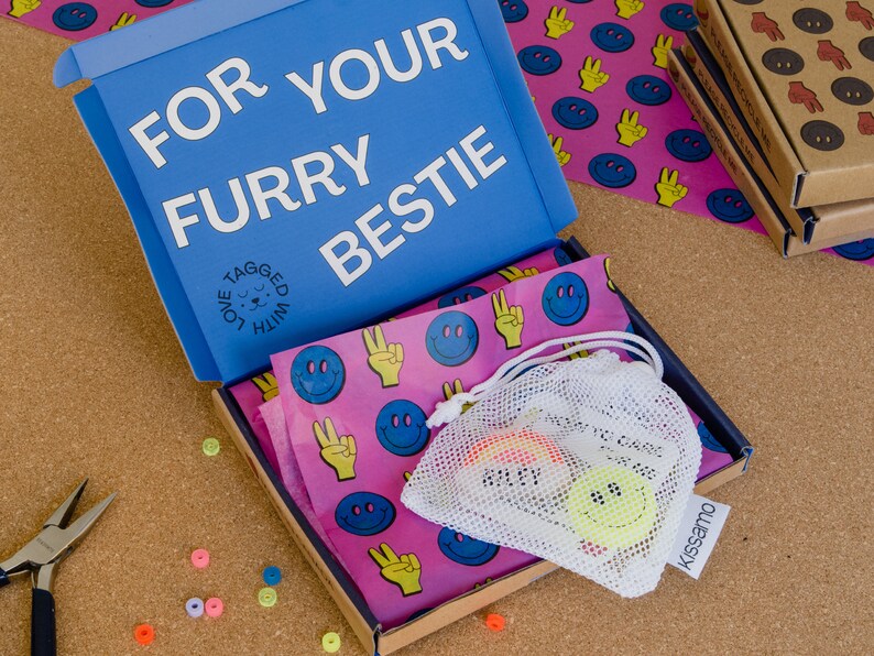 Puede incluir: Una caja azul con el texto "FOR YOUR FURRY BESTIE" y una peque&ntilde;a bolsa blanca con una carita sonriente amarilla en su interior. La caja est&aacute; abierta y el interior est&aacute; forrado con papel rosa con caritas sonrientes azules y signos de paz amarillos.
