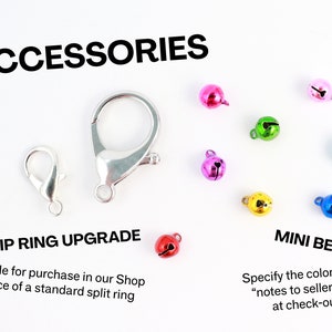 Puede incluir: Primer plano de varios accesorios de plata y metal para la elaboraci&oacute;n de joyas. La imagen muestra dos tipos diferentes de cierres, un anillo de clip de plata y una variedad de peque&ntilde;as campanas de diferentes colores. El texto "ACCESSORIES" est&aacute; en la parte superior de la imagen. El texto "CLIP RING UPGRADE" est&aacute; debajo de la imagen del anillo de clip. El texto "MINI BELL" est&aacute; debajo de la imagen de las campanas. El texto "Available for purchase in our Shop in place of a standard split ring" est&aacute; debajo del texto "CLIP RING UPGRADE". El texto "Specify the colour in the "notes to seller" box at check-out" est&aacute; debajo del texto "MINI BELL".