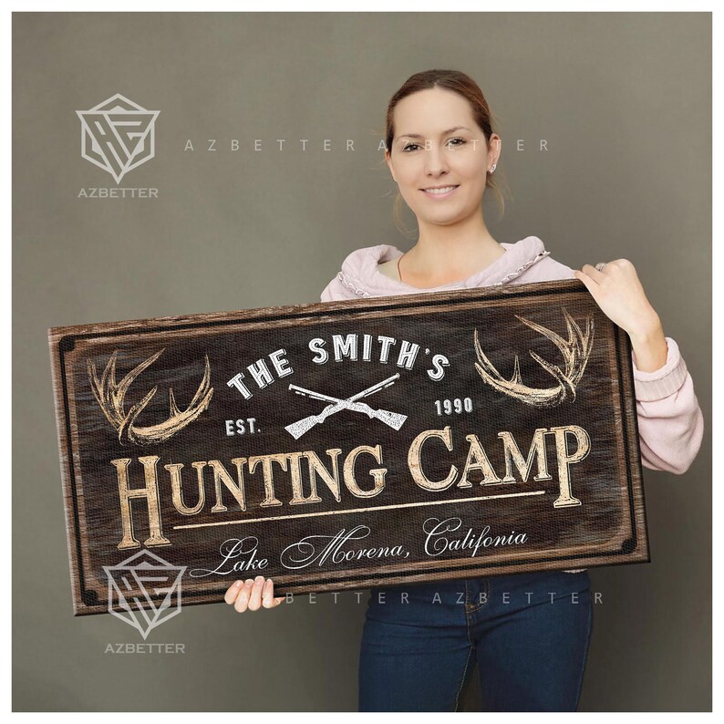 May include: A rustic wooden sign with the text "THE SMITH'S HUNTING CAMP" and "Lake Morena, California." The sign features crossed rifles, antlers, and the year 1990. The sign is brown with white and tan lettering.