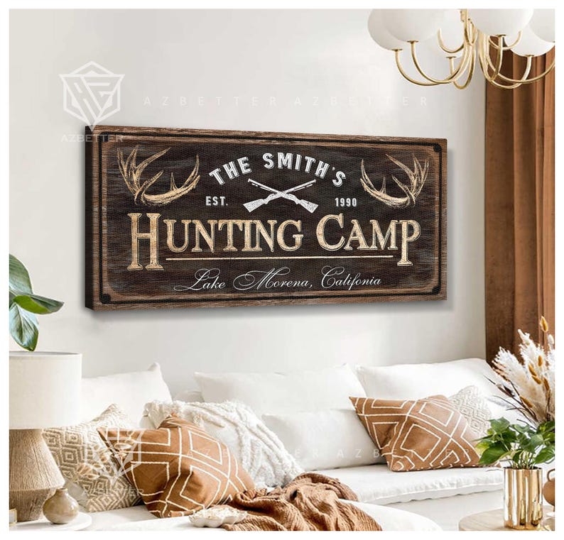 May include: A rectangular wooden sign with the text "THE SMITH'S HUNTING CAMP" in a rustic style. The sign includes crossed rifles, antlers, and the text "Lake Morena, California". The sign is mounted on a wall above a white couch.